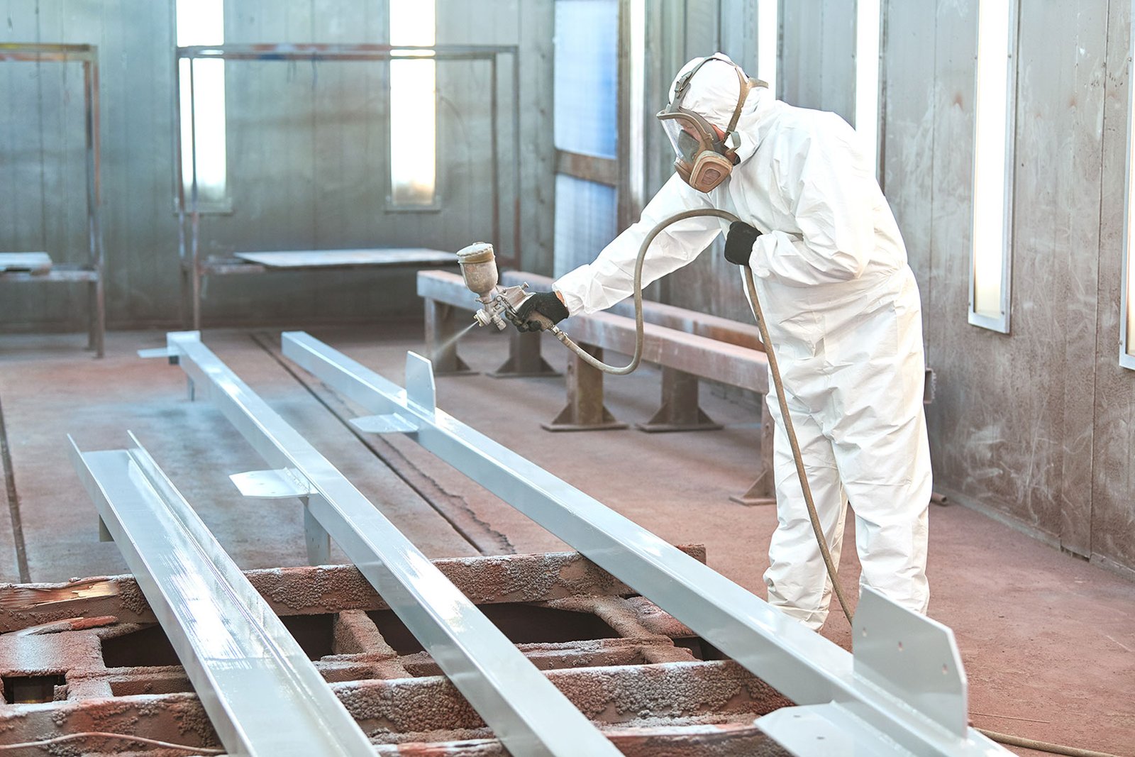 Person in protective suit and respirator spray-painting long metal beams in an industrial facility.